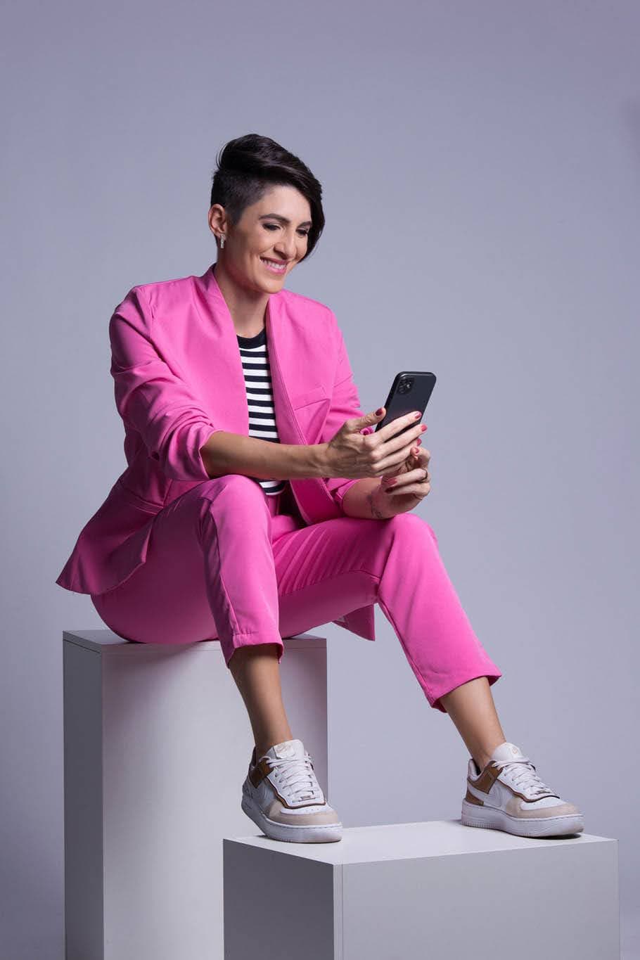 Larissa Zorzenon de terno rosa, sentada, sorrindo enquanto olha o celular